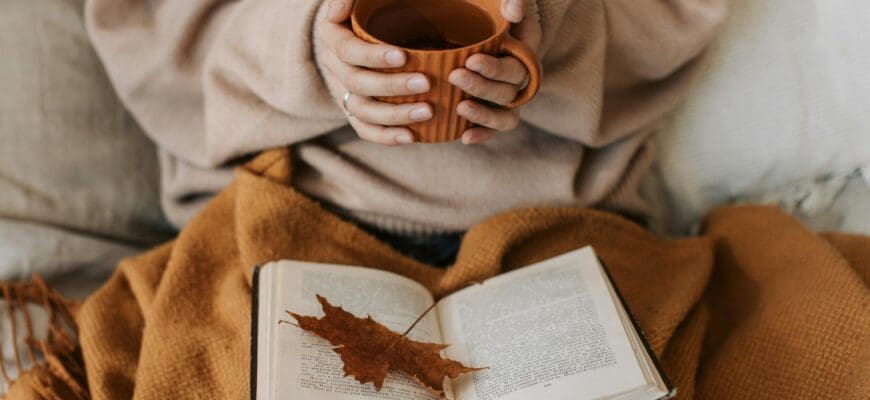 Woman reading a book with a coffee in hand under a brown blanket